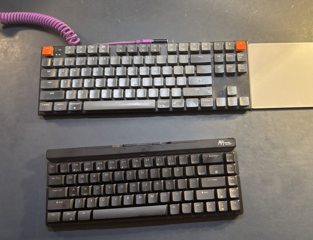 Two mechanical keyboards placed on a dark surface. The top keyboard has colorful keycaps, while the bottom keyboard has a more minimalist design.